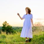Beautiful woman in white dress hat with bouquet of flowers on meadow, in sunset